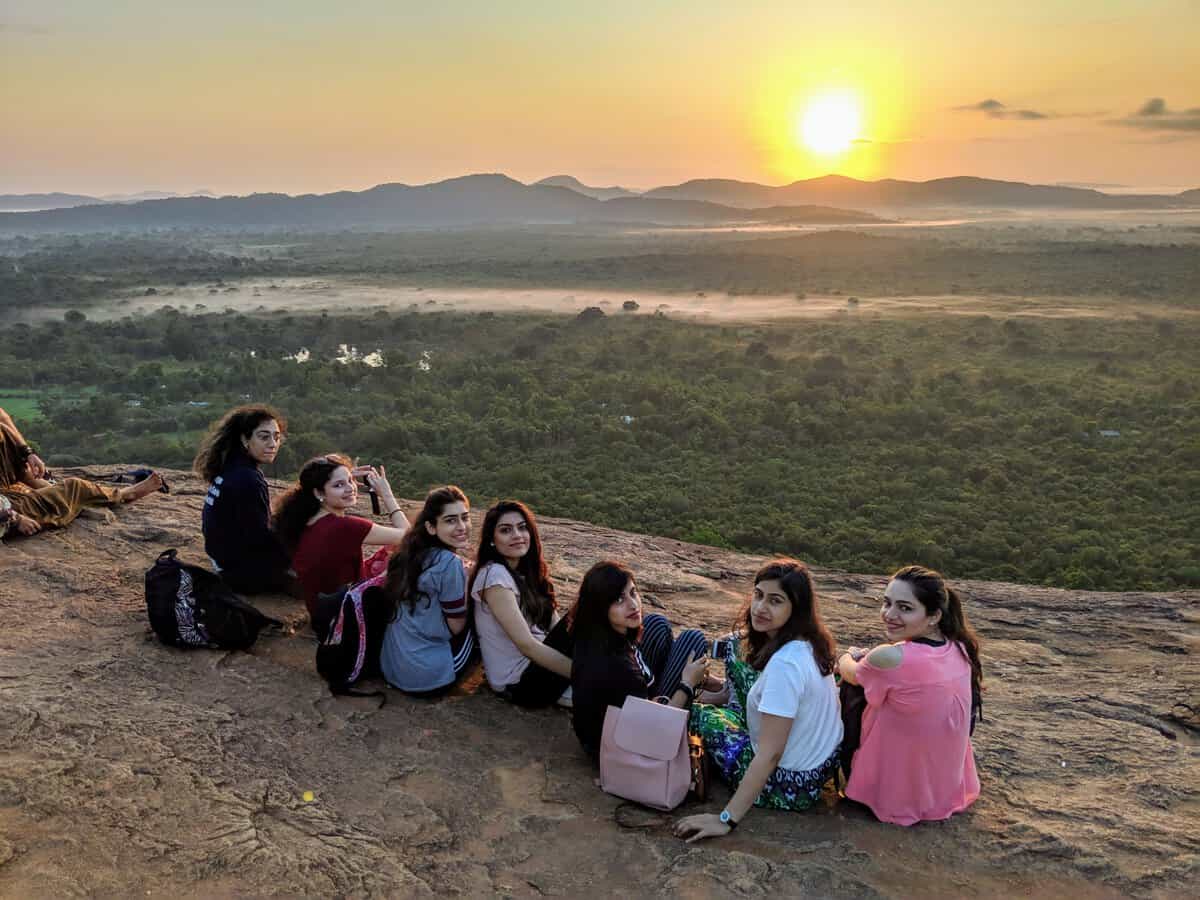 A group of women stands together on the rocky summit of Pidurangala at sunrise, overlooking the surrounding forests and distant hills. The early morning light and sweeping views capture the rewarding experience of completing the popular sunrise hike.