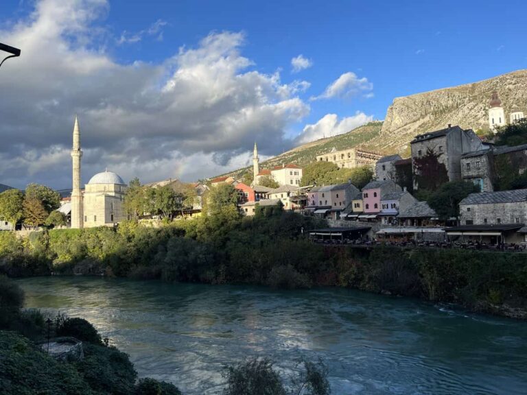 View of the Neretva River in Mostar with the Apashi Mosque in the distance, framed by trees and stone buildings, under a cloudy sky. The scene captures the calm, layered beauty of the city’s east side.