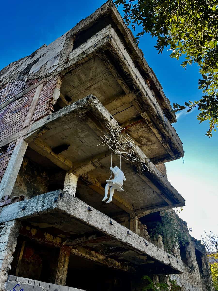 Street art memorial in Mostar showing a painted child hanging from a swing attached to an abandoned building. The artwork is stark and emotional, contrasting innocence with the building’s war-damaged facade.