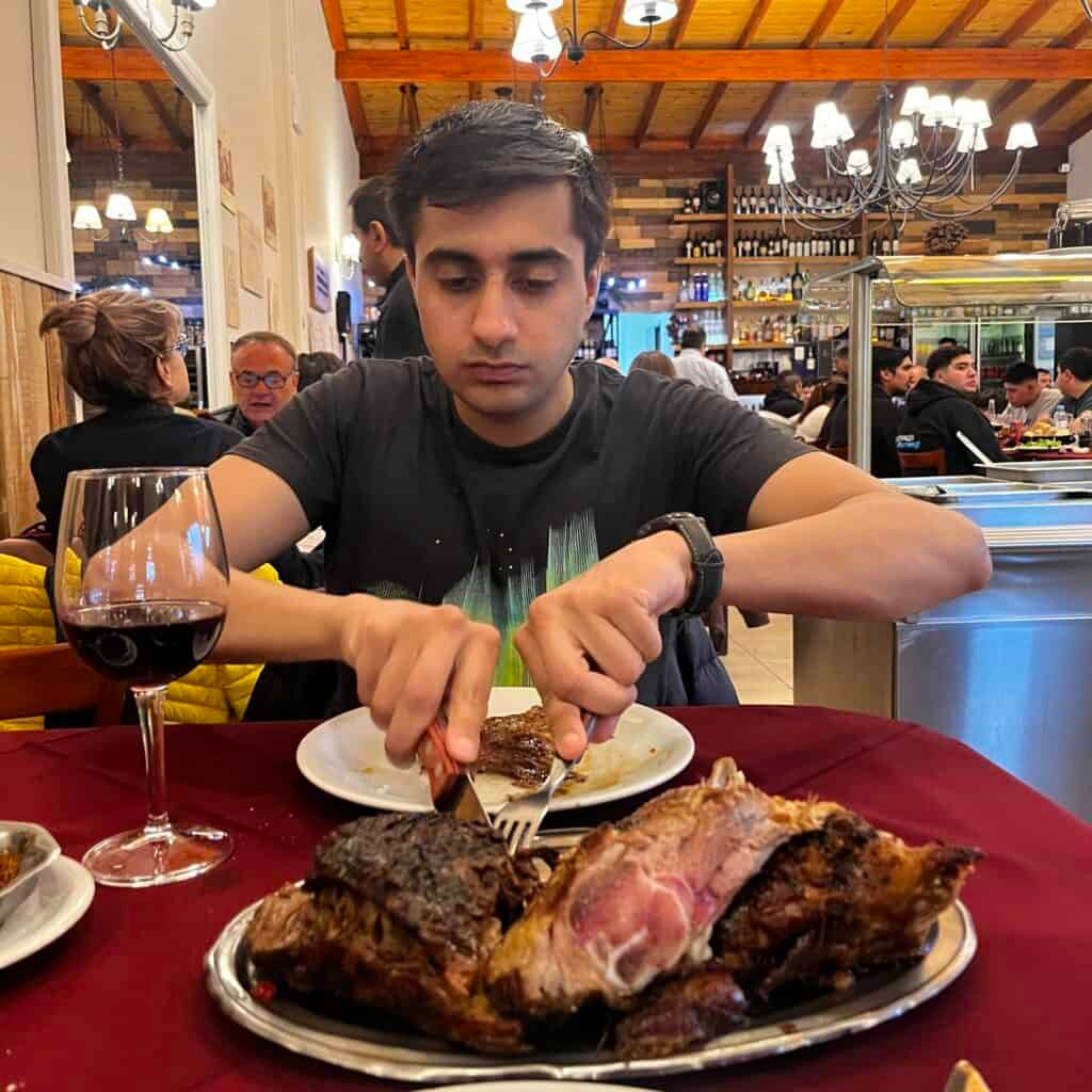 A man cuts into a thick grilled steak served on a plate at a restaurant in Ushuaia. The meal showcases the hearty meat dishes commonly enjoyed in Patagonia.
