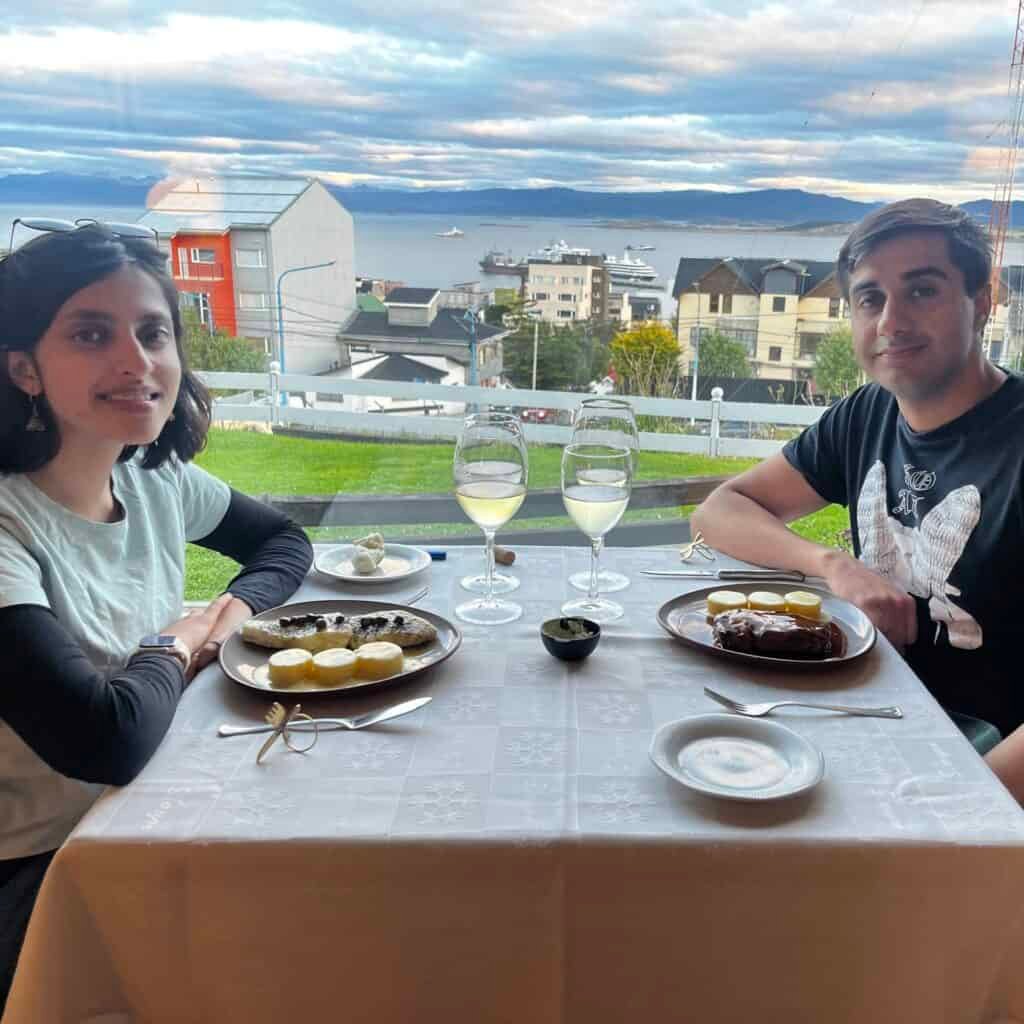 A couple sits together at a restaurant in Ushuaia with full plates of food and glasses of white wine on the table. The cozy meal captures the experience of dining out during a trip to Ushuaia.