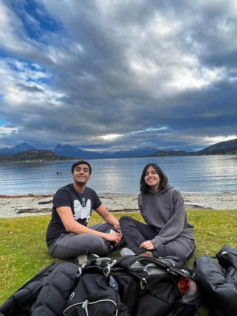 Two people sit together on the grass in Tierra del Fuego National Park with calm blue water stretching out behind them. Forested hills and mountains surround the peaceful Patagonian landscape.