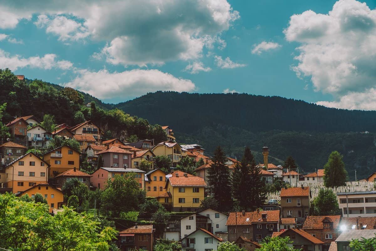 Colorful low-rise buildings spread across the Sarajevo valley, with green hills rising behind them under a clear sky. A peaceful cityscape that captures the layered charm of Sarajevo's old and new neighborhoods.