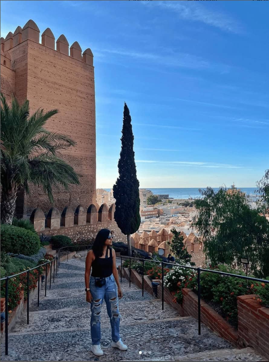 A woman stands mid-step on the stone stairs of a historic castle in Granada, Spain, framed by warm-toned walls and Moorish architectural details. The image complements the home page of Musafir in Transit, highlighting a blend of history and personal journey.