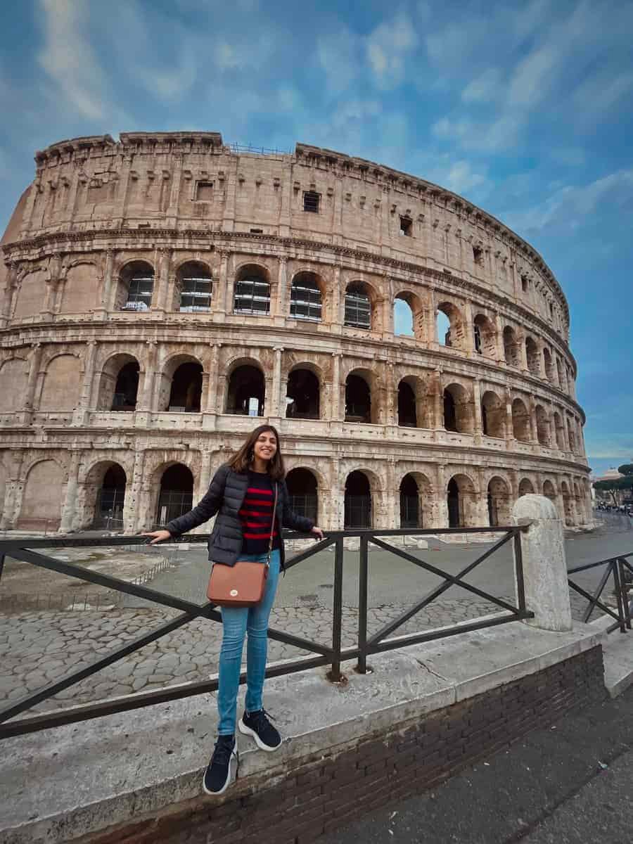 Woman standing in front of the Colosseum in Rome on a sunny morning, smiling with sunglasses and casual travel attire. The ancient amphitheater’s iconic arches rise behind her, capturing the excitement of visiting one of Rome’s most famous landmarks.