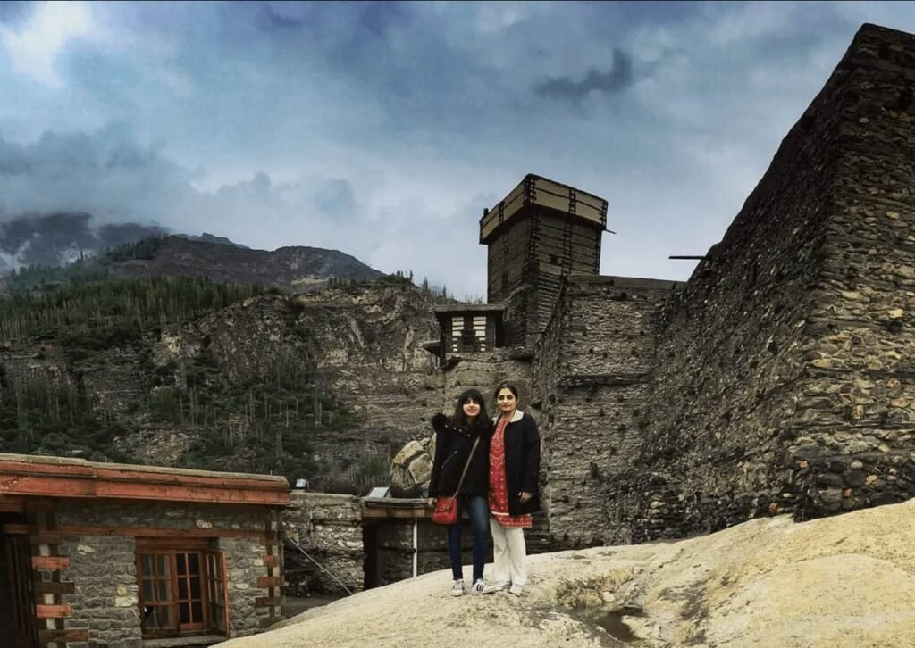 Two women stand in front of the historic Altit Fort in Hunza on a cloudy day. The stone fort and surrounding mountains rise behind them beneath an overcast sky.