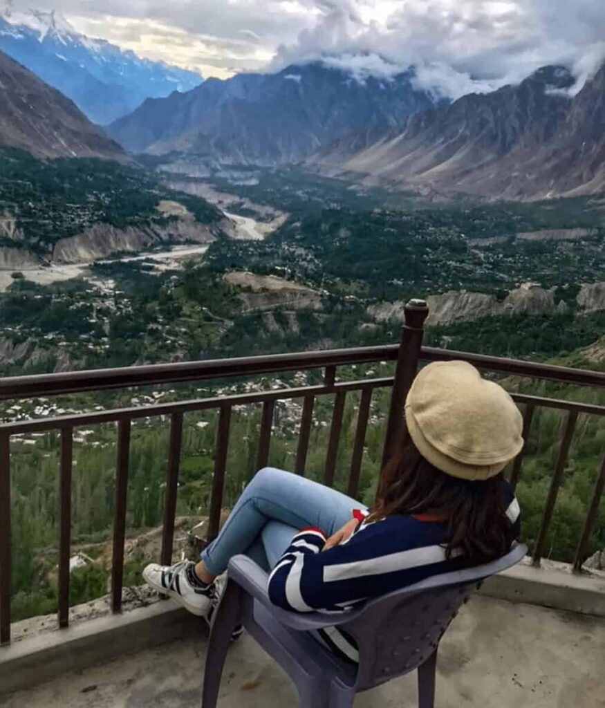 A woman sitting on a balcony facing a valley with mountains in the background.