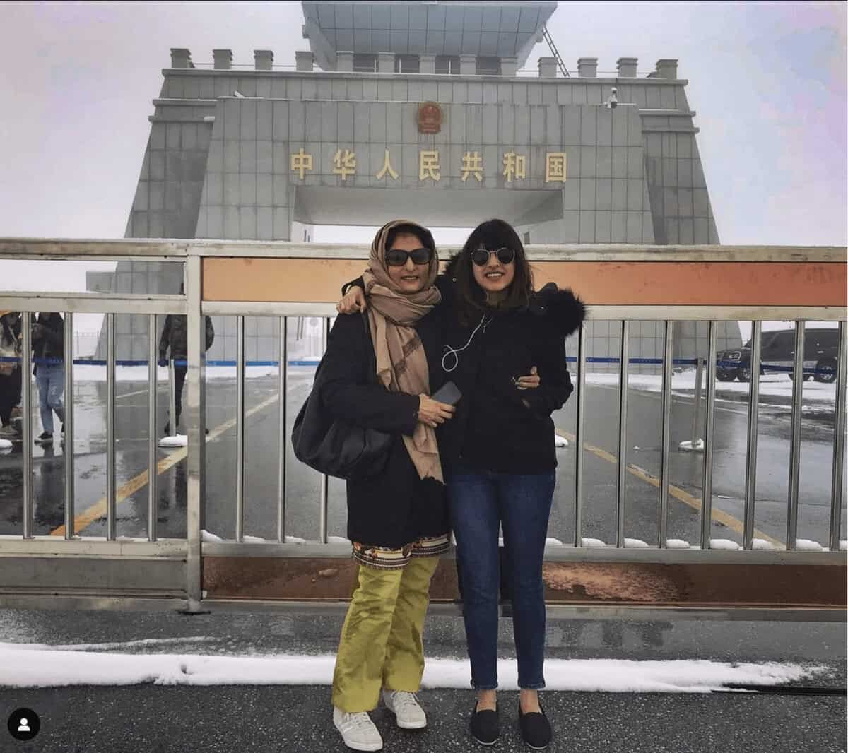 Two travelers stand in front of the Khunjerab Pass monument marking the Pakistan China border. Snow covered mountains surround the high altitude border crossing.