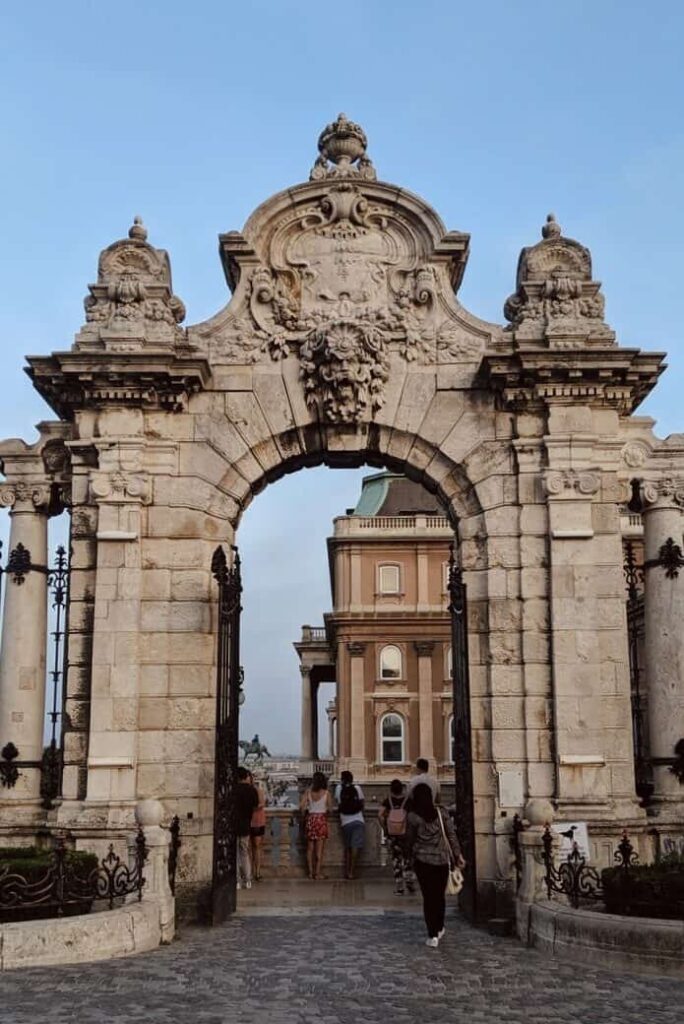 A grand, ornate gate with ironwork detail marks the entrance to Buda Castle, flanked by stone columns and sweeping city views in the background. The gate hints at the historic and royal legacy of the castle complex.