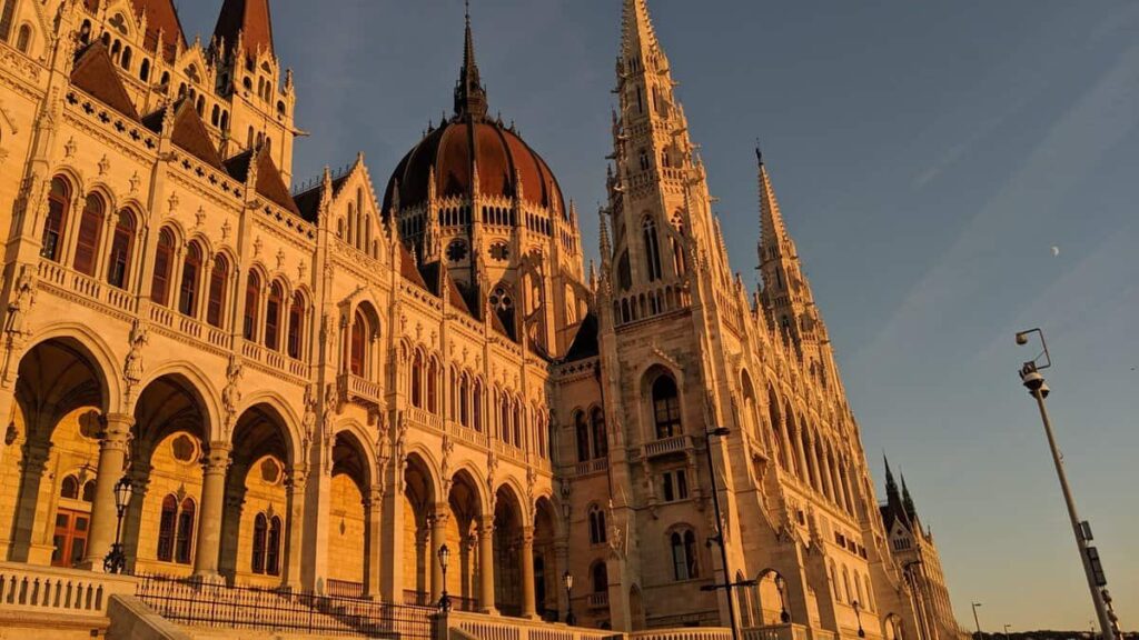 The Hungarian Parliament building glows in warm orange light as the sun sets behind it, with soft pink and gold hues filling the sky. The reflection in the Danube enhances the grandeur of the scene.