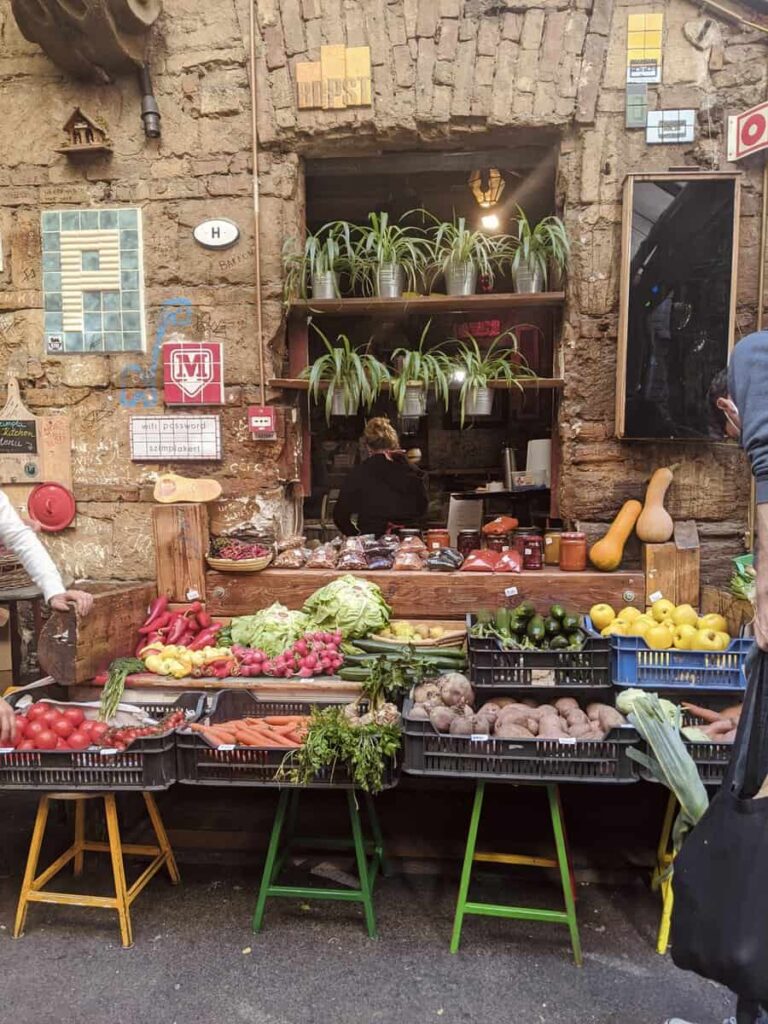 Colorful stalls filled with fresh produce, homemade goods, and flowers line the eclectic interior of Szimpla Kert. Locals and tourists browse casually under string lights and quirky decor.