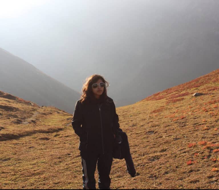 A girl standing in all black, hands in pockets with misty background of mountains