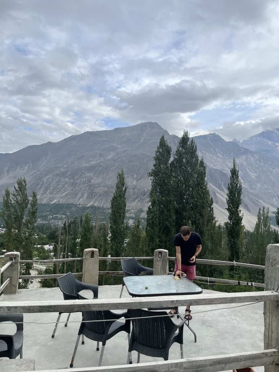 A man standing on a balcony overlooking Hunza Valley, Pakistan, with terraced fields and dramatic mountain ranges stretching into the distance. The view captures the vastness and serenity of the Hunza landscape from an elevated vantage point.