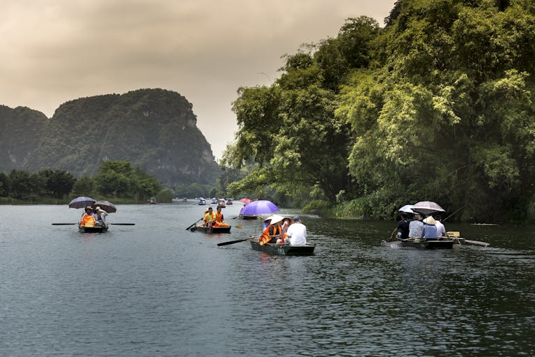 People enjoy a serene boat ride on a calm river surrounded by lush trees and distant mountains.