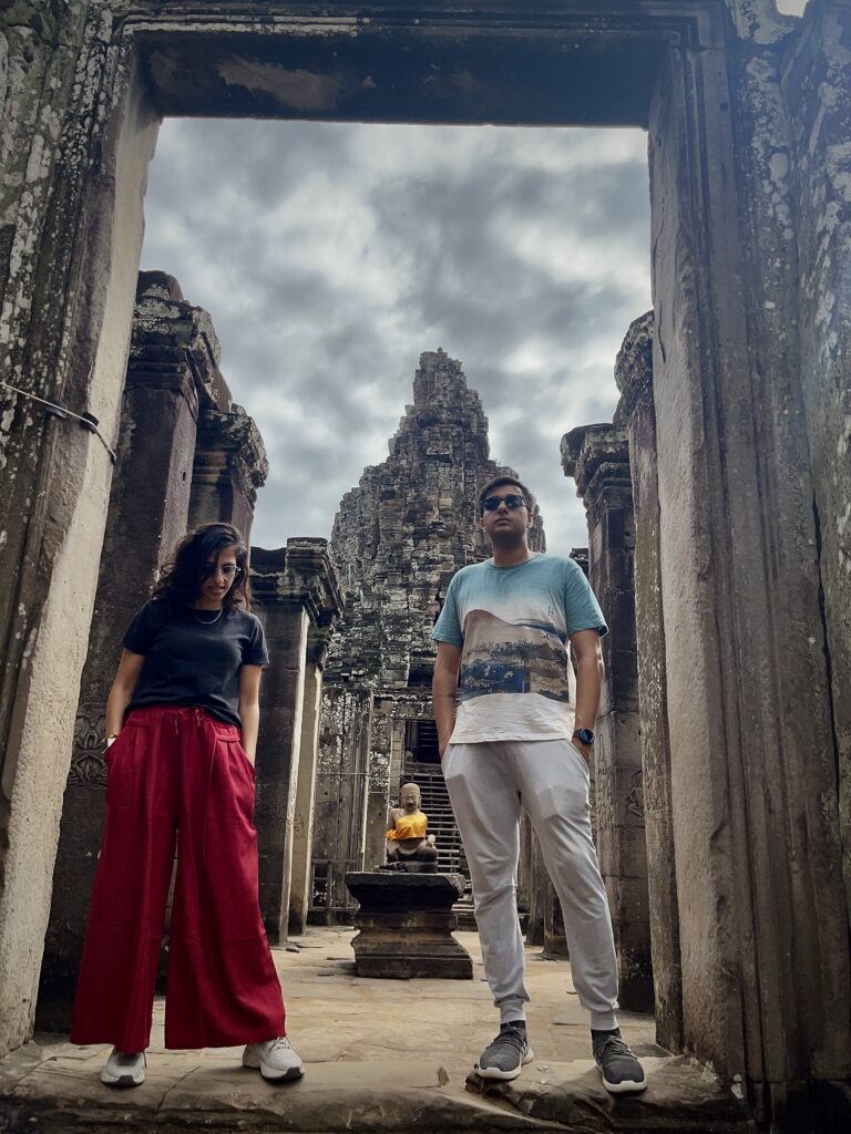 Two people stand in the doorway of an ancient temple in Siem Reap, Cambodia, with the iconic stone faces of Bayon visible in the background. The temple’s weathered stone and intricate carvings frame the figures.