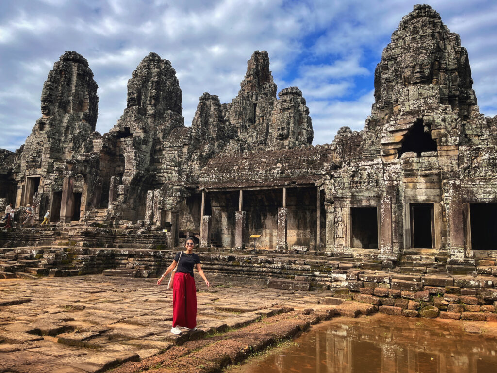 Zoomed-out view of a person standing at Bayon Temple in Siem Reap, Cambodia. The temple’s iconic stone towers with carved faces rise in the background, highlighting the grandeur and intricate detail of this ancient Khmer architecture.