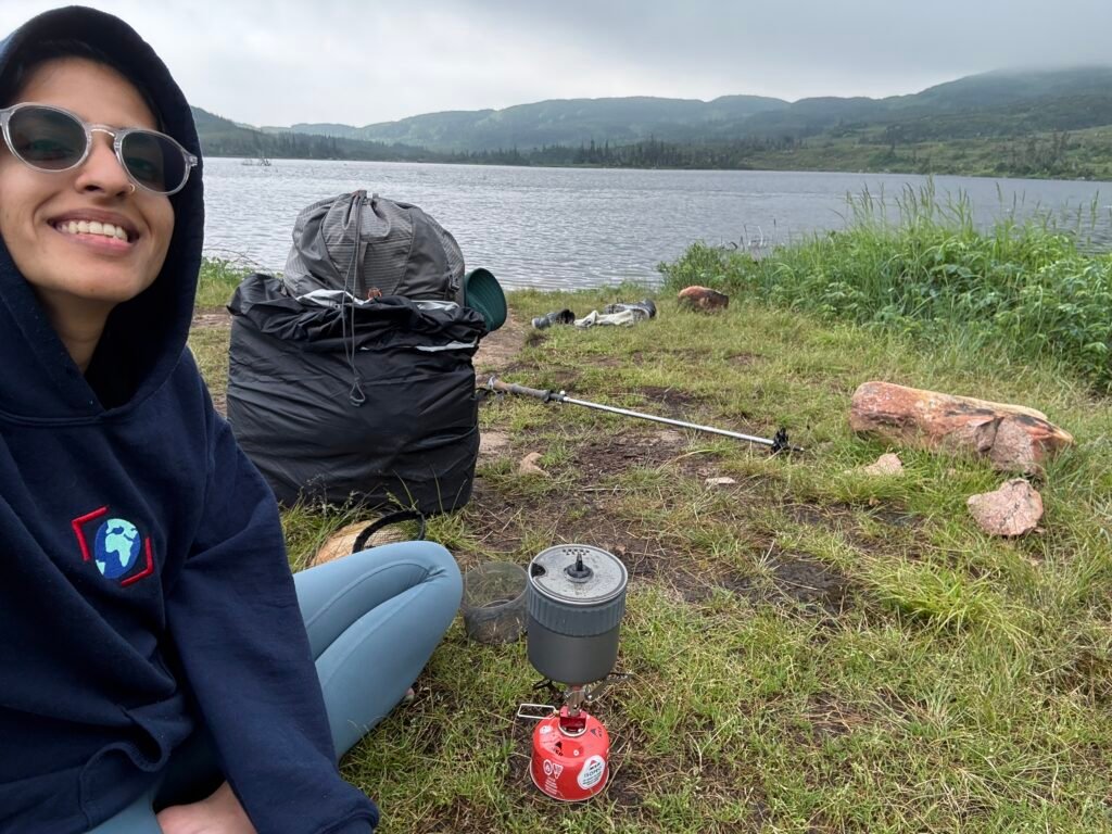 Selifie taken by a woman next to a lake, infront of her is a small stove the picture depicts her about to cook a meal