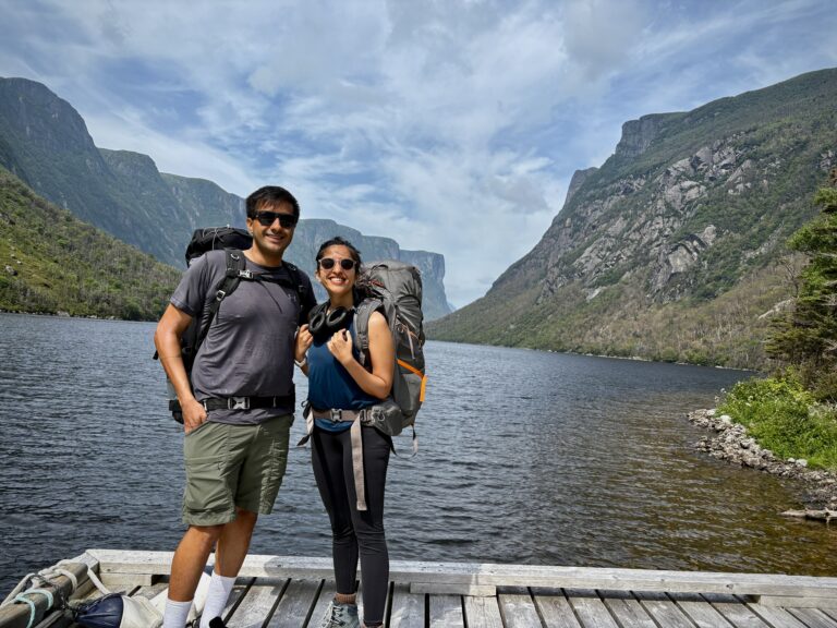 A couple with loaded backpacks stands on the dock at Western Brook Pond, smiling and ready to begin the Long Range Traverse. The calm water and steep fjord walls in the background hint at the remote hike ahead.