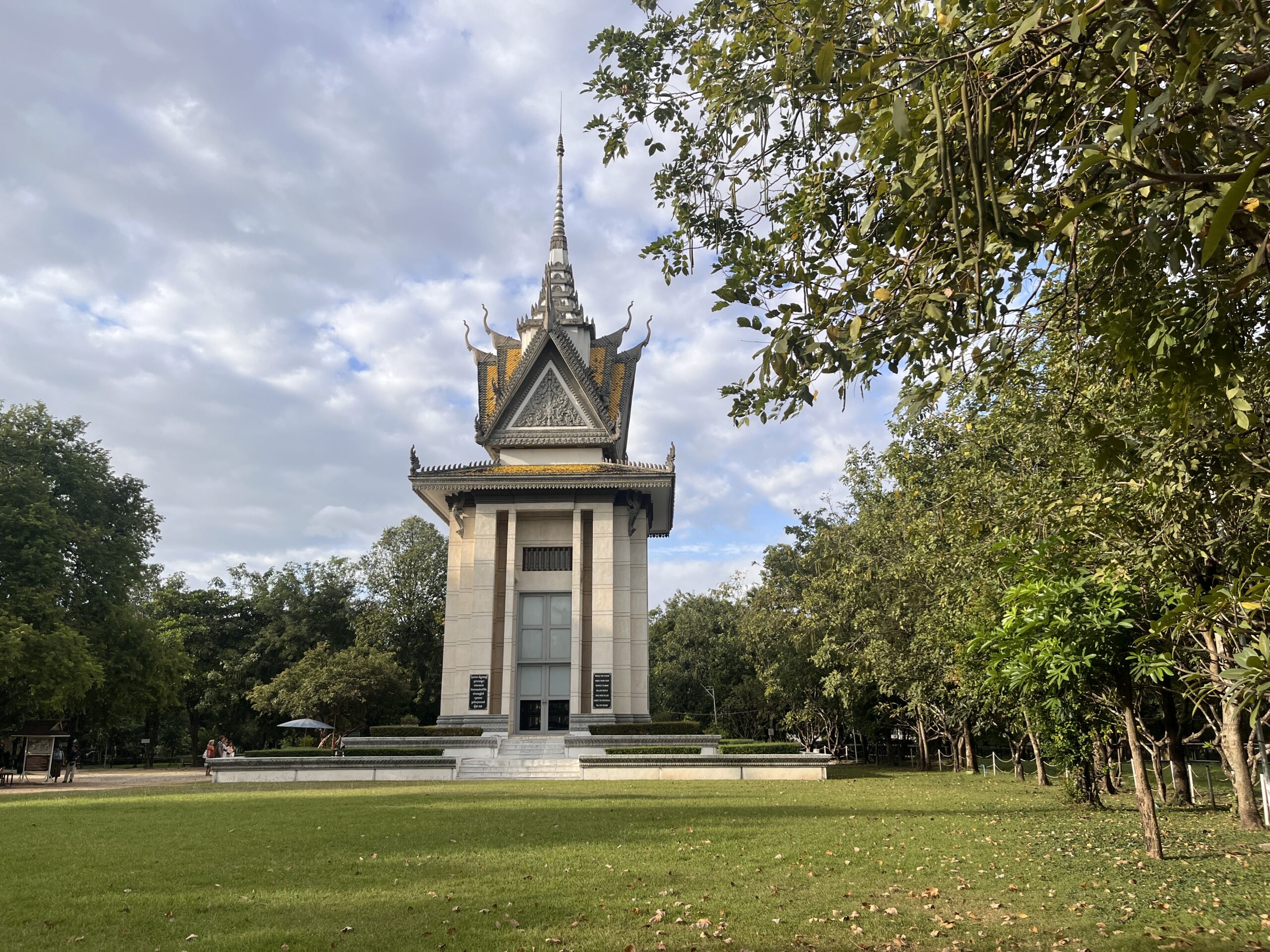 Distant view of the memorial stupa at the Killing Fields in Cambodia, housing the skulls of genocide victims. The glass structure rises solemnly against the sky, symbolizing remembrance and the scale of the tragedy.