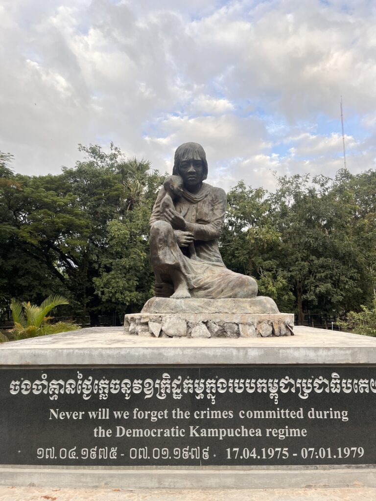 Statue of a mother holding a child at the Killing Fields memorial in Cambodia, with the words “Never Forget” inscribed below. The sculpture conveys deep grief and remembrance, honoring the victims of the Khmer Rouge regime.