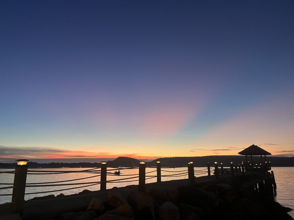 Sunset view from a beach on Koh Rong Samloem, Cambodia, with warm orange and pink hues lighting the sky. A wooden pier stretches into the calm sea, creating a serene and tropical island atmosphere.