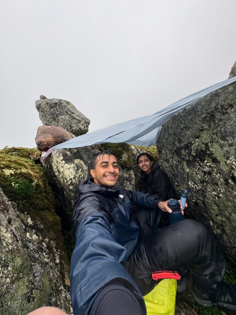 A couple takes a selfie while sheltering from heavy rain under a picnic blanket stretched between two large rocks, weighted down with another rock. They’re wedged in a rocky crevice, smiling in full rain gear during a downpour on the Long Range Traverse.