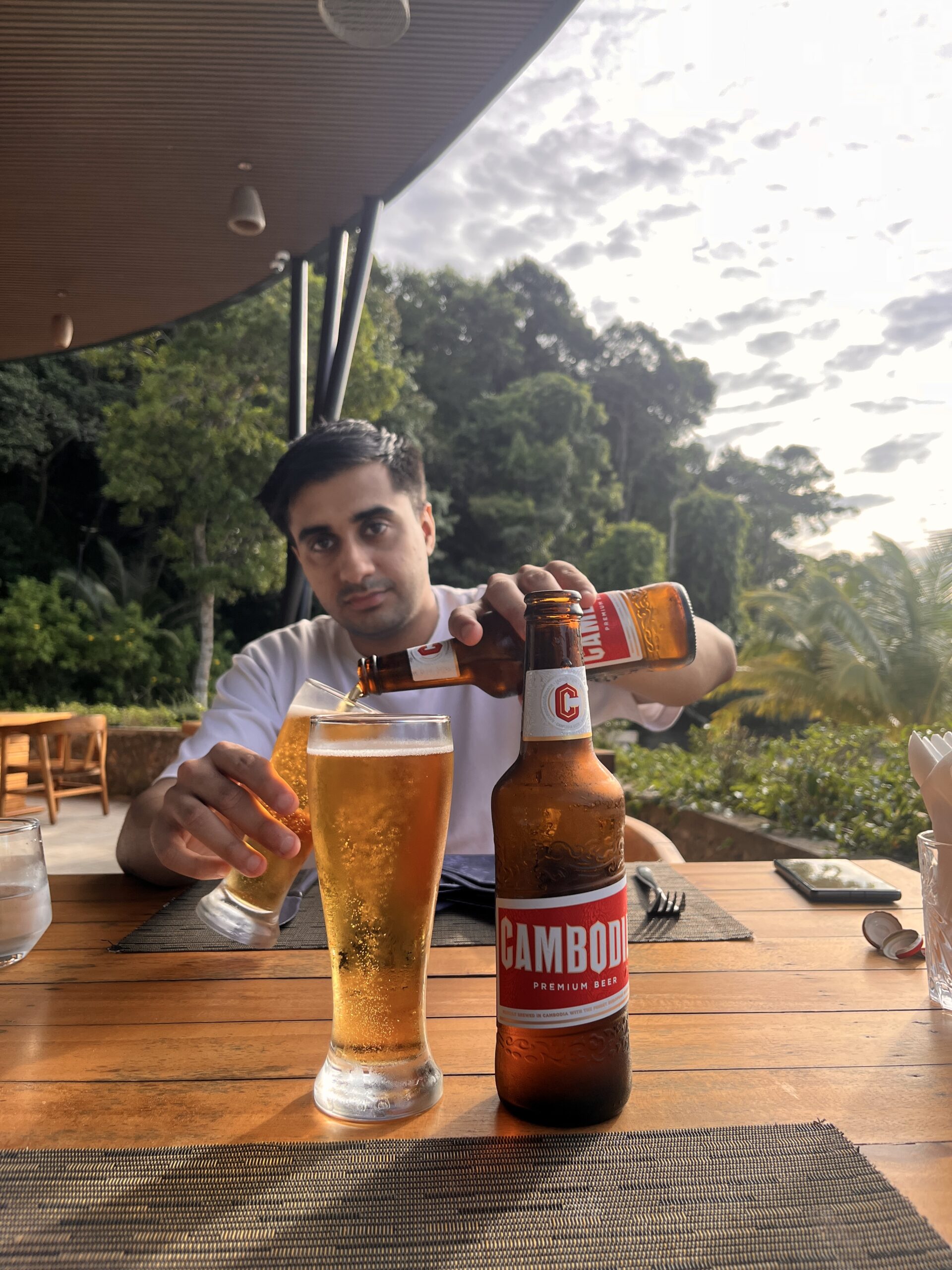 A man pours Cambodia Premium Beer into a chilled glass at an outdoor restaurant table surrounded by lush greenery on Koh Rong Samloem, with soft afternoon light filtering through the trees.
