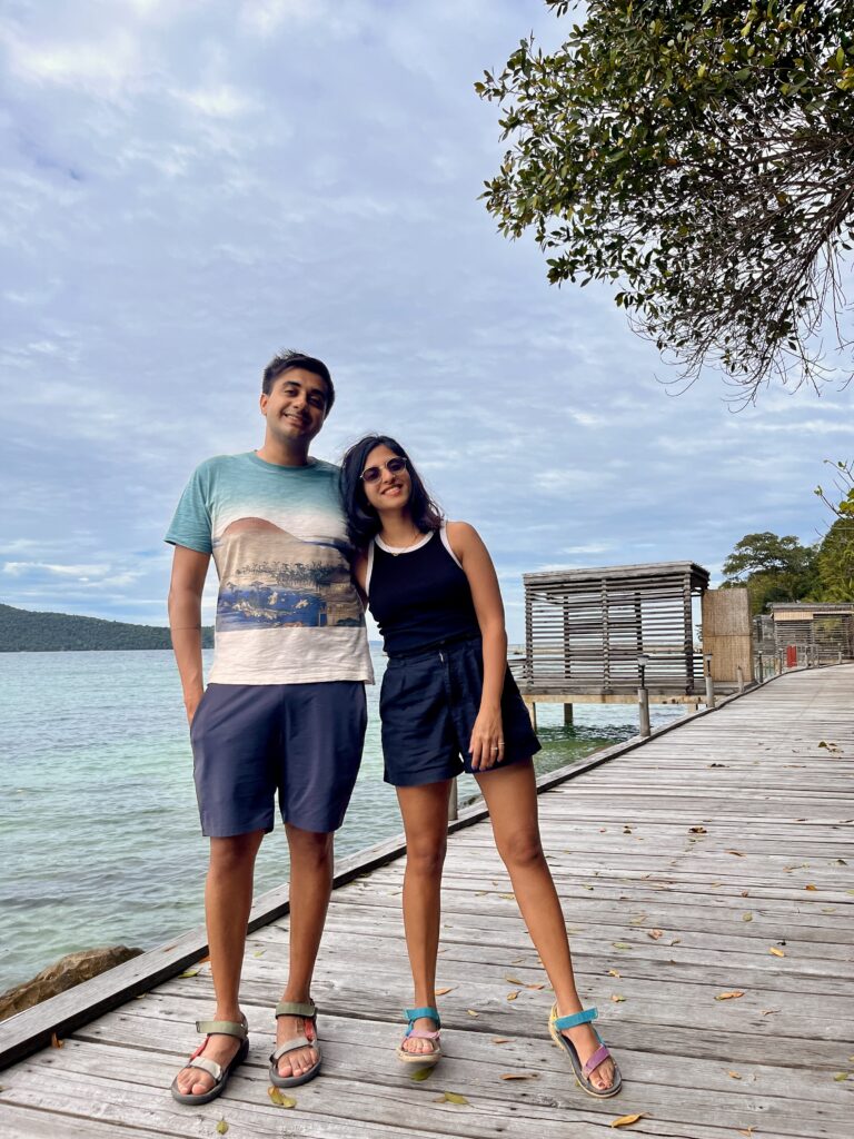 A smiling couple stands arm-in-arm on a wooden boardwalk by the turquoise water on Koh Rong Samloem, with rustic beach huts and forested hills in the background under a cloudy sky.