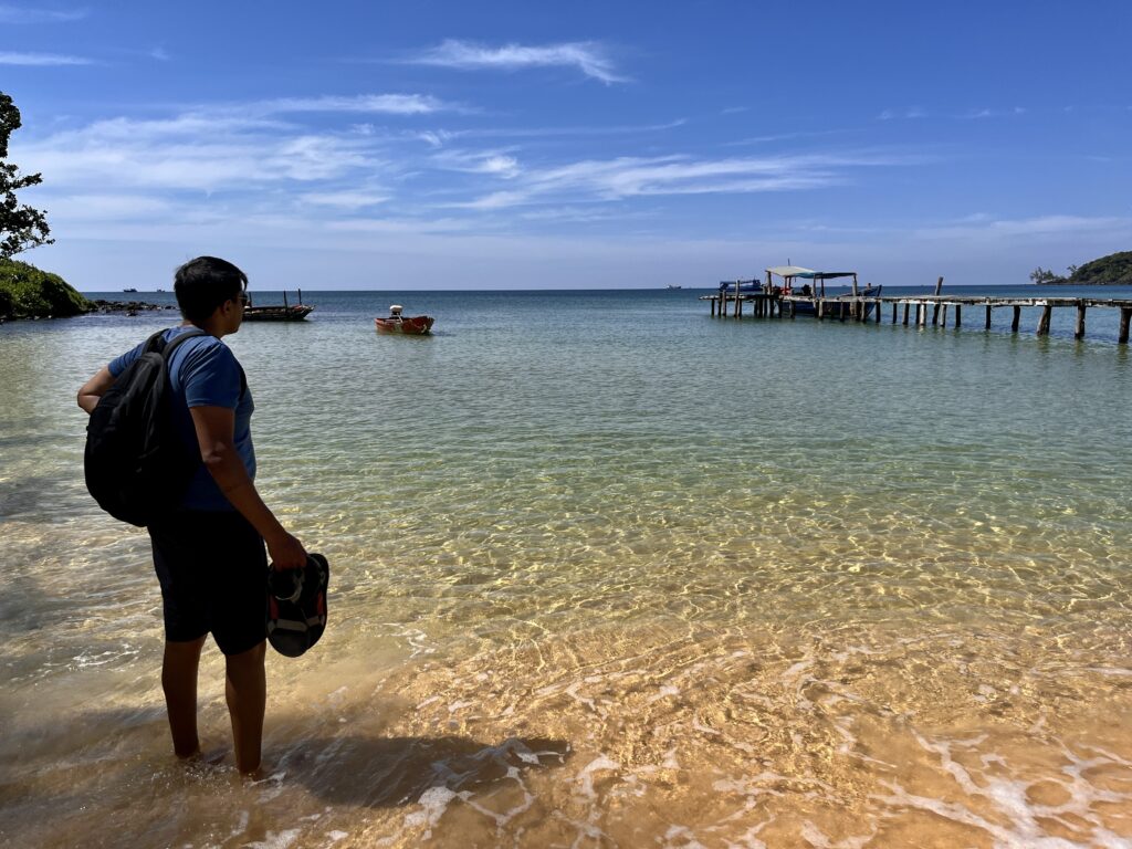 A man stands ankle-deep in the clear turquoise water of Koh Rong Samloem, holding his sandals and looking out toward wooden fishing boats and a weathered pier under a bright blue sky. The golden sand and gentle waves create a tranquil, tropical scene.