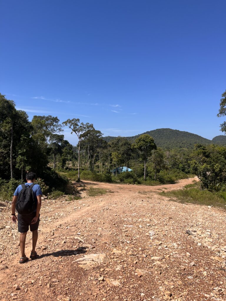 A man wearing a backpack walks down a rocky red dirt trail surrounded by dense jungle and distant hills under a clear blue sky on Koh Rong Samloem.