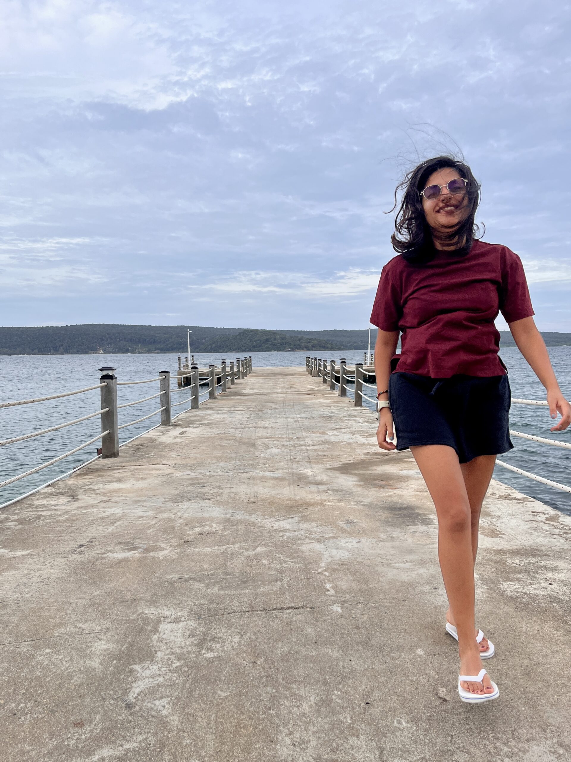A woman in a burgundy t-shirt and black shorts walks barefoot on a concrete pier with the sea on both sides, smiling as the wind blows through her hair on a cloudy day in Koh Rong Samloem.