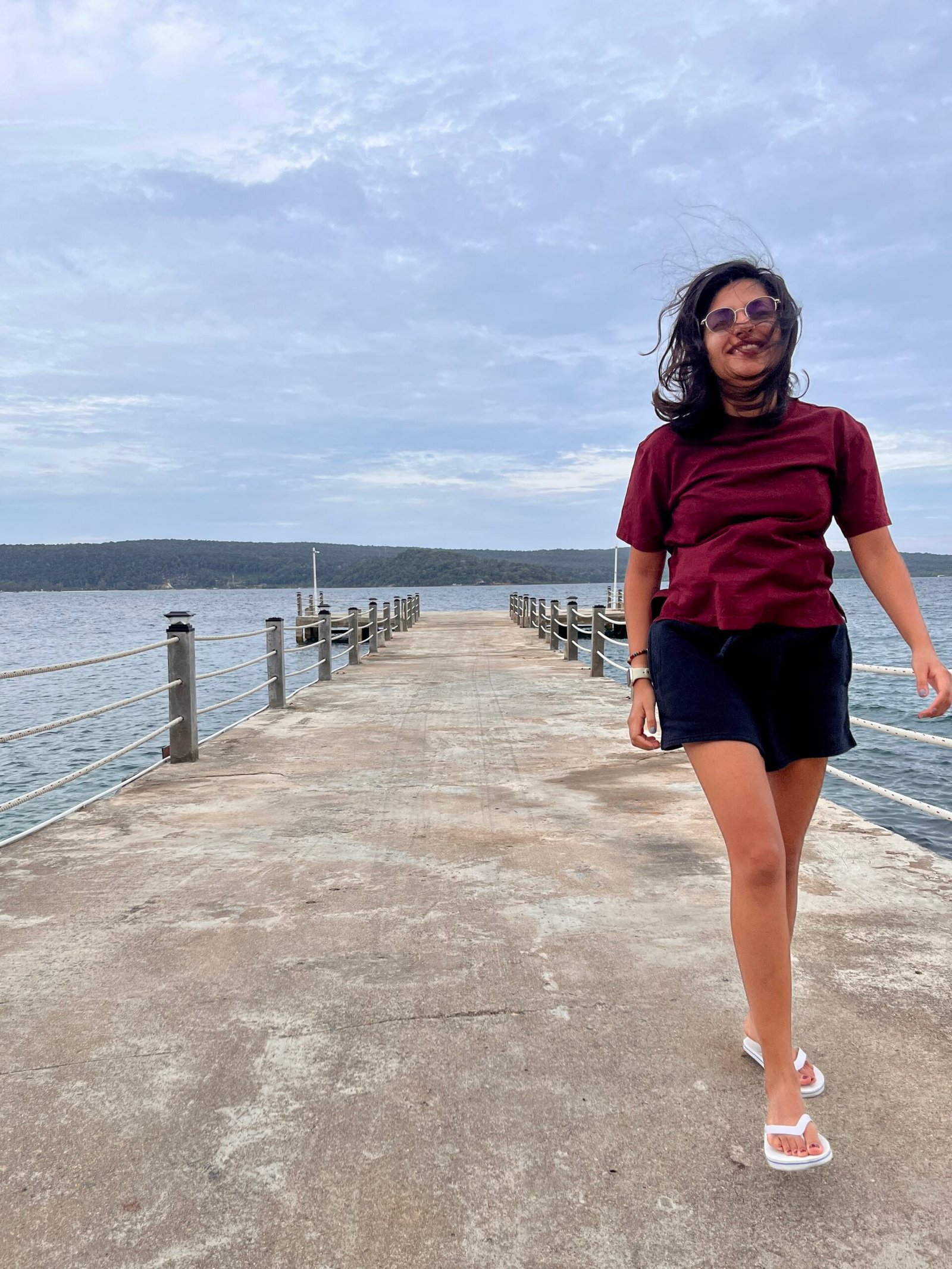 A woman in a burgundy t-shirt and black shorts walks barefoot on a concrete pier with the sea on both sides, smiling as the wind blows through her hair on a cloudy day in Koh Rong Samloem.