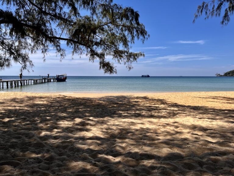 Shaded golden sand stretches toward the calm turquoise sea at Lazy Beach, Koh Rong Samloem, with a wooden pier jutting into the water and fishing boats anchored nearby under a clear blue sky. Pine tree branches frame the scene, casting dappled shadows over the sand.