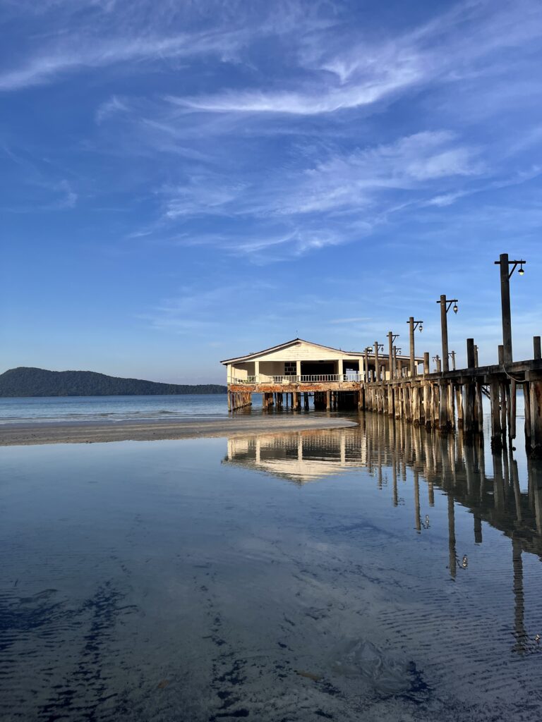 A wooden pier with a rustic beachfront building extends over shallow, glassy water in Sacren Bay, Koh Rong Samloem, reflecting the structure and blue sky in the still surface below.