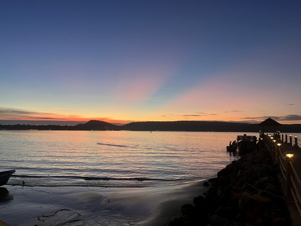 A vivid sunset casts warm orange and purple hues across the sky and water at Pearl Beach Resort and Spa in Koh Rong Samloem, with a softly lit pier extending into the sea and silhouetted hills lining the horizon. Gentle waves lap at the sandy shore, adding to the serene atmosphere.