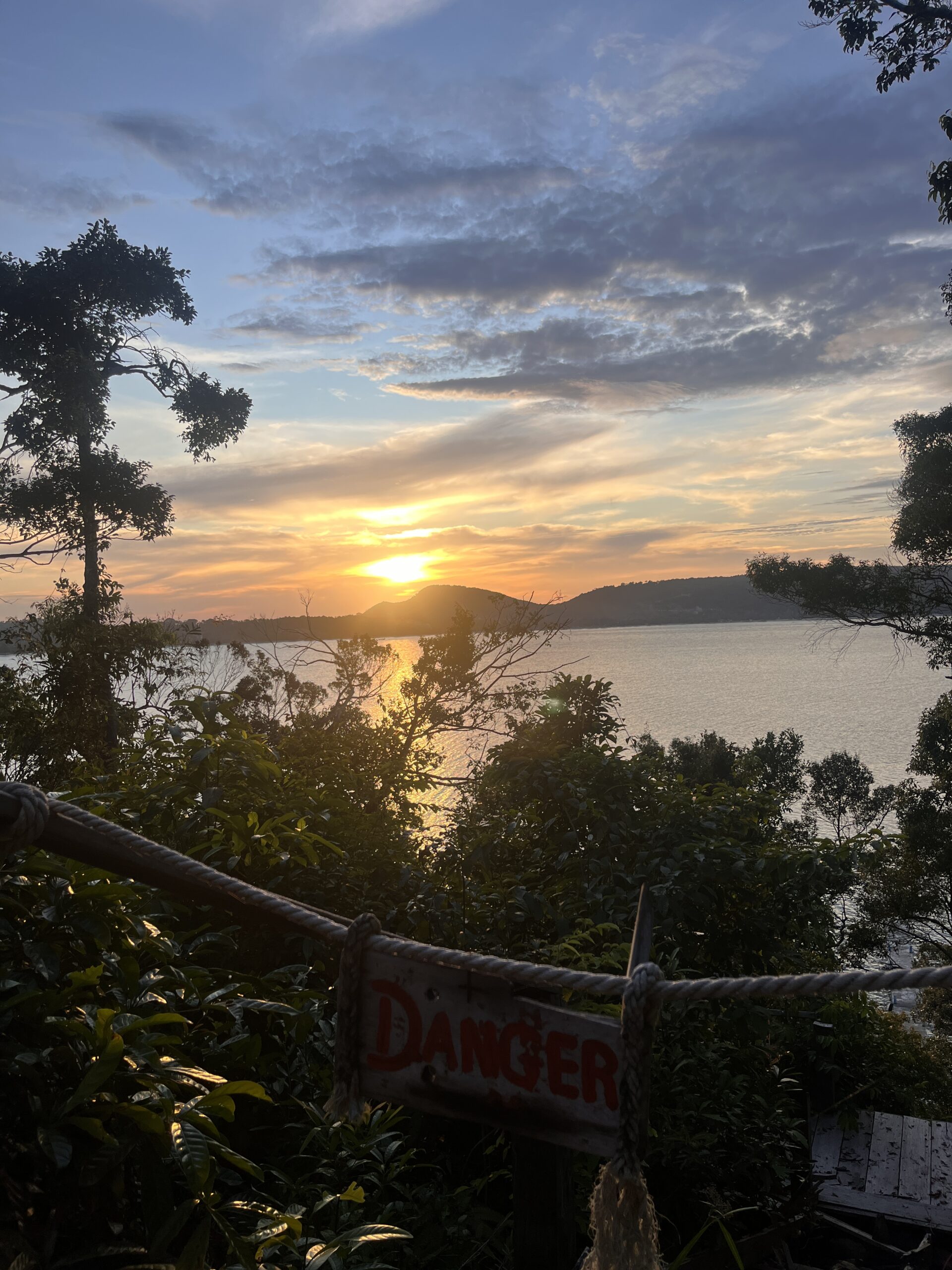 A vivid sunset over the ocean seen through dense greenery on Koh Rong Samloem, with a red "Danger" sign tied to a rope in the foreground marking the edge of a cliff or steep drop.