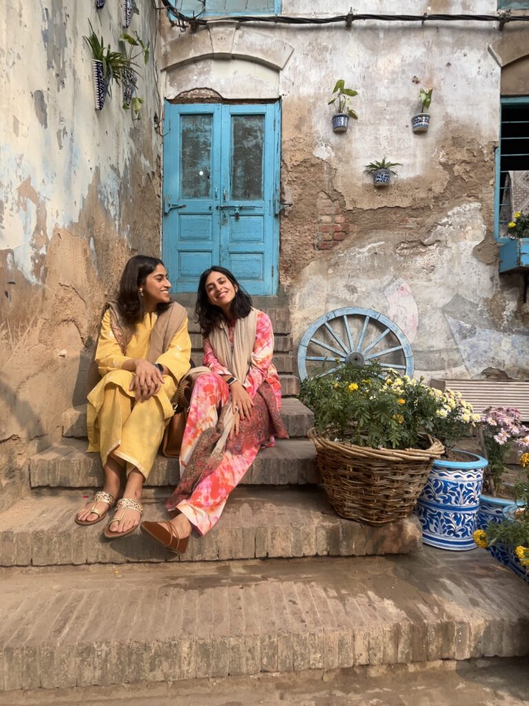 Two women in colorful traditional clothes sit on sunlit brick steps in a rustic Old Lahore courtyard, surrounded by vibrant flowers, blue planters, and a distressed wall with hanging pots.