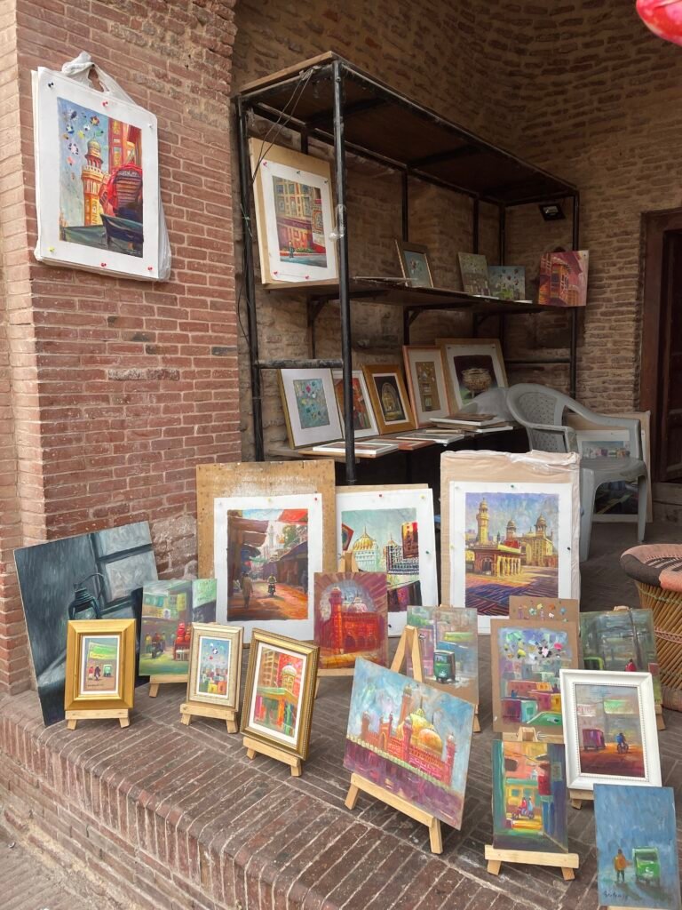 A street art display in Old Lahore featuring colorful framed paintings of iconic landmarks and everyday scenes, arranged against rustic brick walls near a heritage site.
