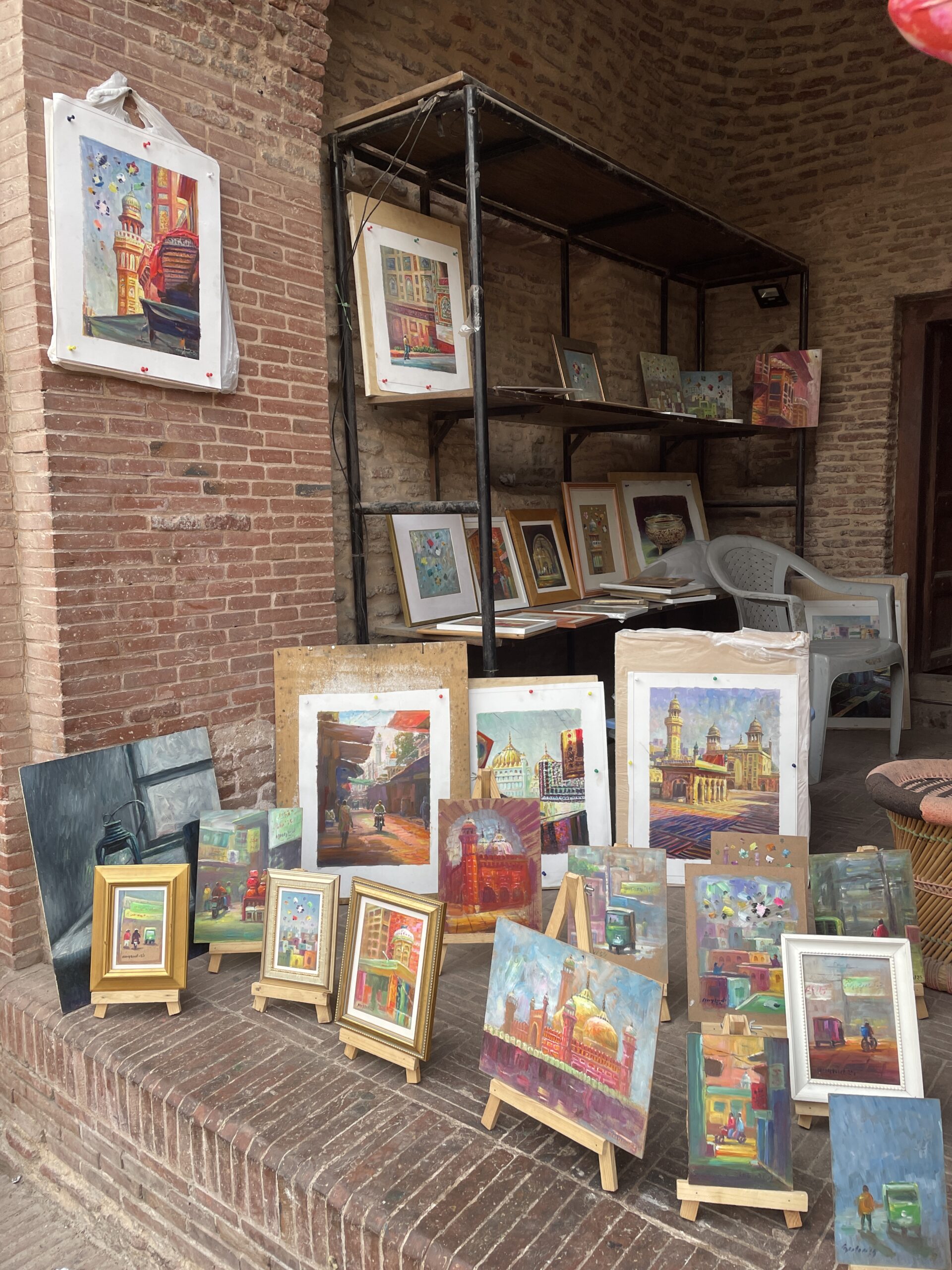A street art display in Old Lahore featuring colorful framed paintings of iconic landmarks and everyday scenes, arranged against rustic brick walls near a heritage site.