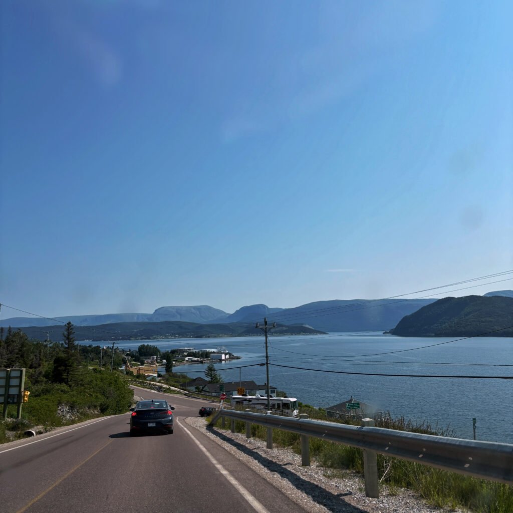 Alt Text A car drives along a winding coastal highway in Newfoundland, with calm blue water on one side and distant hills under a clear summer sky. The scenic drive captures the feeling of starting a road trip west from St. John’s.