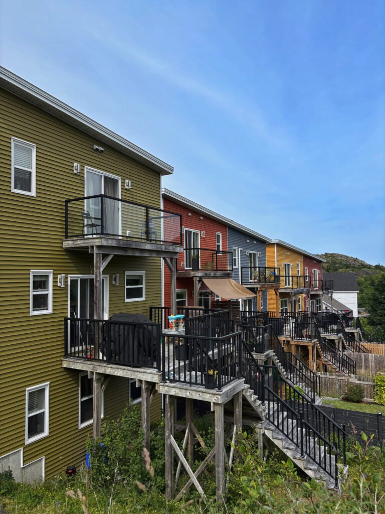 A row of brightly painted hillside homes in St. John’s, Newfoundland, with wooden staircases and back decks overlooking the slope. The houses are painted in shades of green, red, blue, and yellow, capturing the charm of the city’s famous Jellybean Row.