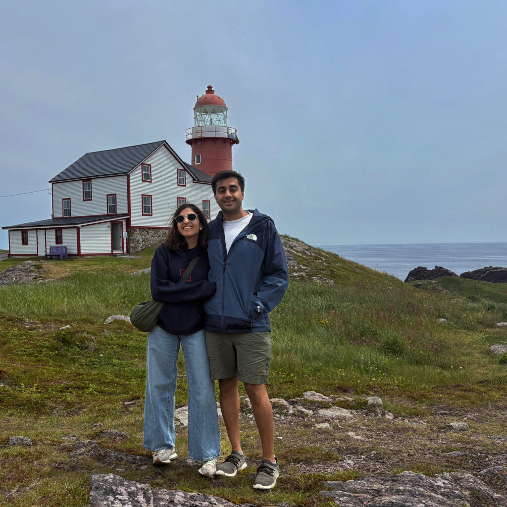 A couple stands on a grassy hill in front of the Cape Spear Lighthouse, with the Atlantic Ocean stretching out behind it. The lighthouse’s red-and-white tower rises above the cliffs, marking the easternmost point in North America.