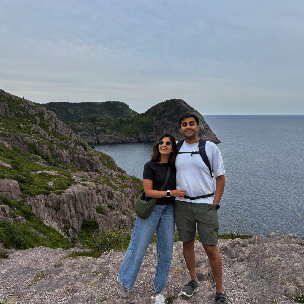 A couple stands on a rocky trail at Signal Hill with dramatic cliffs and the Atlantic Ocean in the background. Surrounded by rugged terrain and lush greenery, the scene captures the beauty and coastal views of this popular hike in St. John’s.
