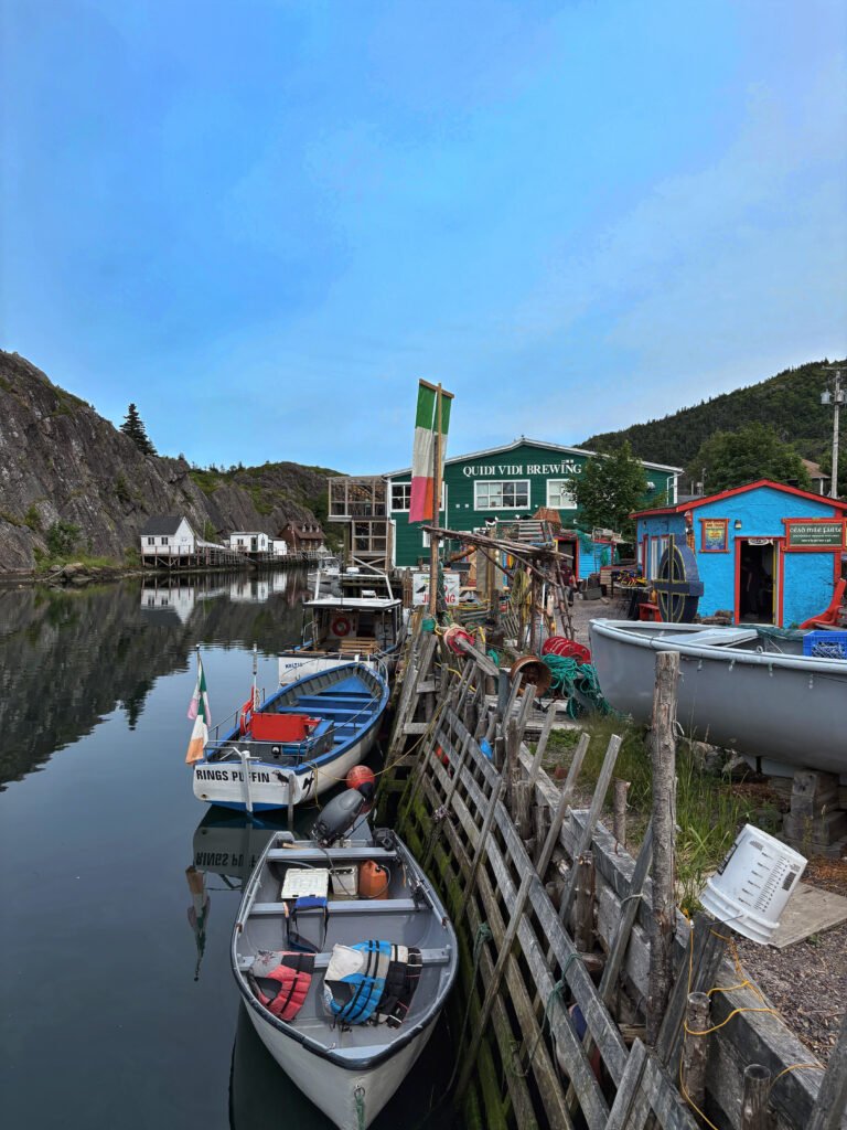 Small fishing boats are tied up along a wooden dock in Quidi Vidi, with colorful sheds, coiled ropes, and the green Quidi Vidi Brewery building in the background. The calm harbor and rugged cliffs frame this lively corner of St. John’s, known for its mix of fishing heritage and craft beer culture.