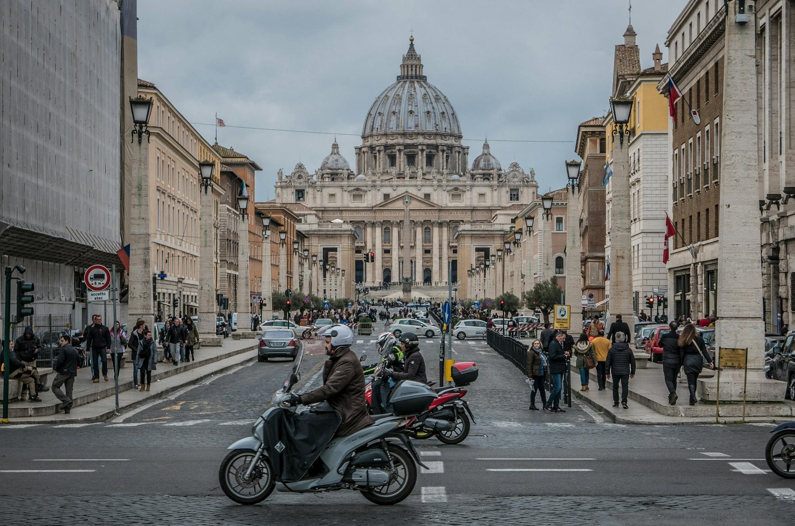 View of St. Peter's Basilica in the distance framed by a lively Roman street filled with people, cafes, and warm afternoon light.