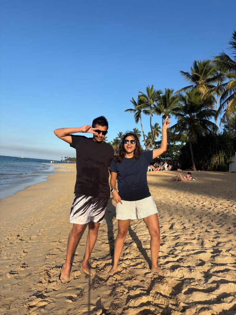 A couple stands barefoot on the sandy beach in Las Terrenas, smiling and posing with palm trees and calm ocean water behind them.