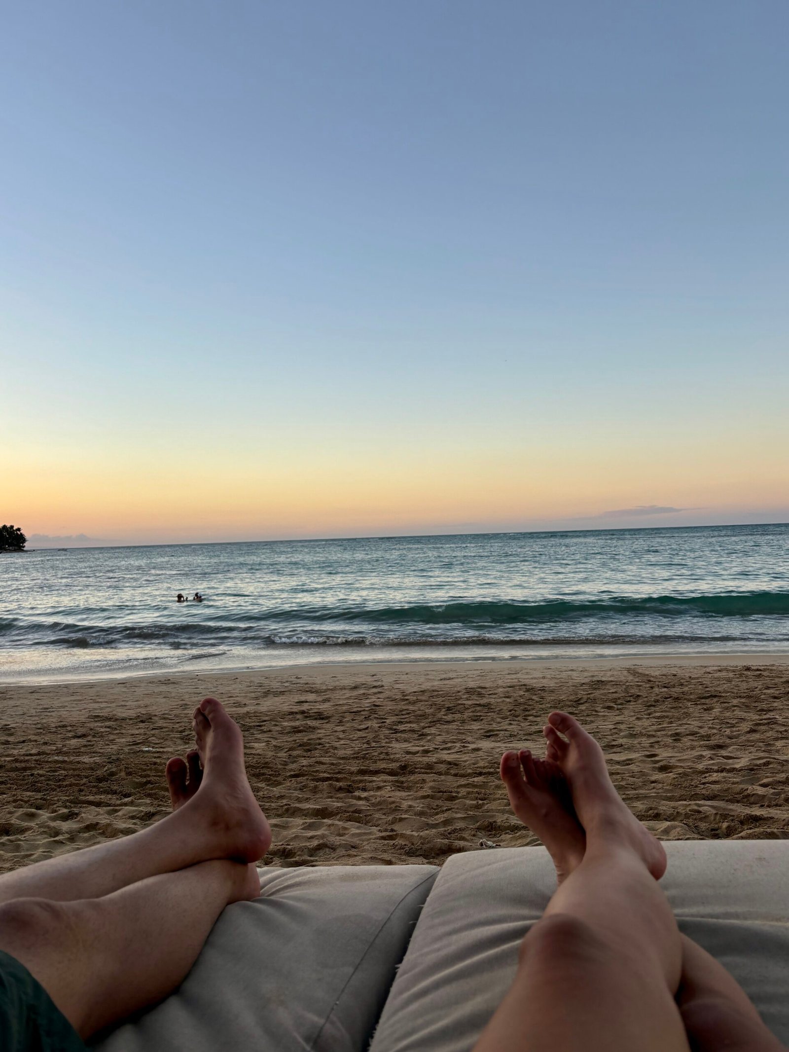 Two people relax on cushioned loungers facing the ocean at sunset in Las Terrenas. The sky fades from soft orange to blue while gentle waves roll onto the sandy shore, creating a calm and unhurried evening beach atmosphere.