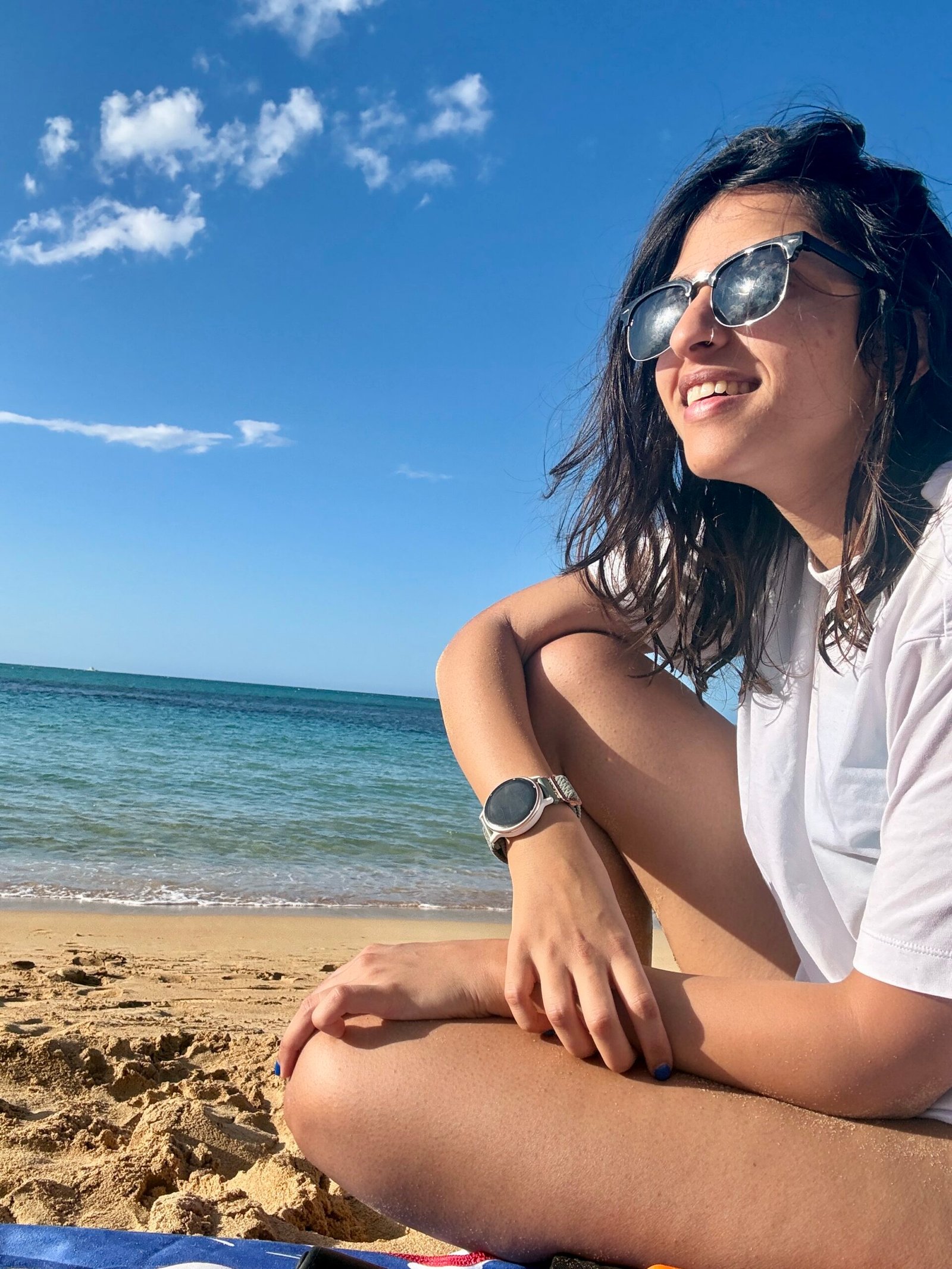 A woman sits on golden sand near the shoreline in Las Terrenas, wearing sunglasses. There is the ocean under a bright blue sky behind her. The clear turquoise water and relaxed posture capture the easy pace of beach life in town.