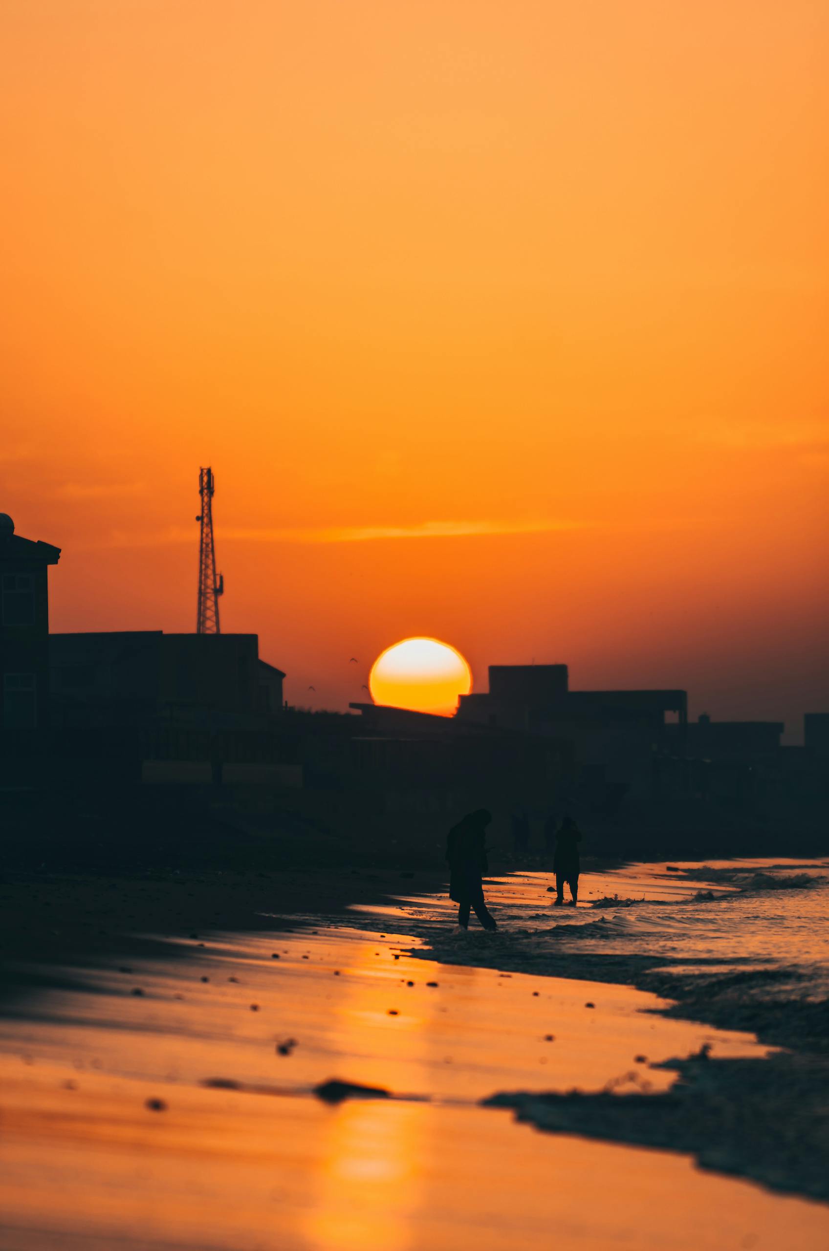 Silhouette of people walking along Karachi beach at sunset with vibrant orange sky.