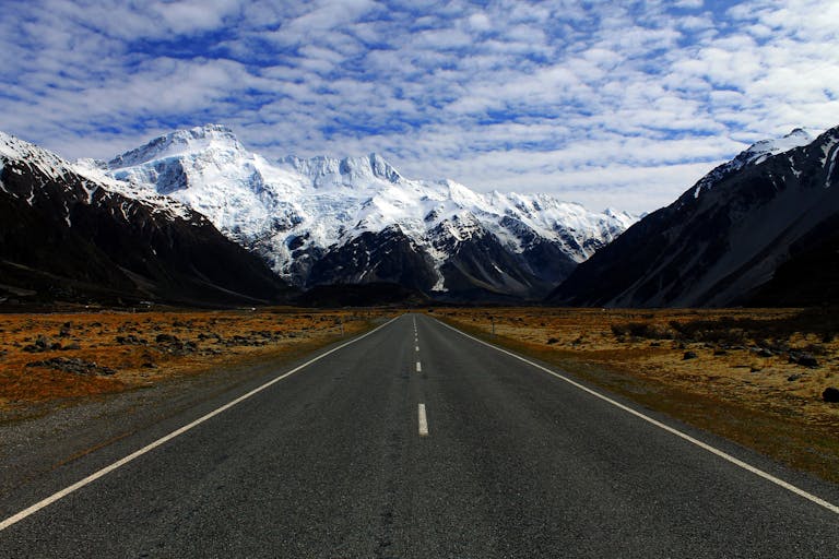 A deserted road leading to majestic snow-covered mountains under a cloudy blue sky.